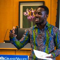 man smiling in front of a podium, holding a paper, "Grand Valley State University" on a plaque behind him